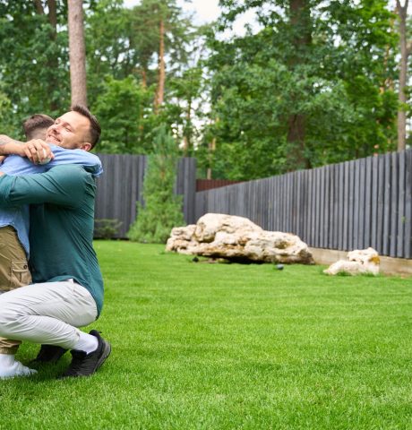 Smiling father hugs his son on the lawn of the house, the boy has a baseball glove